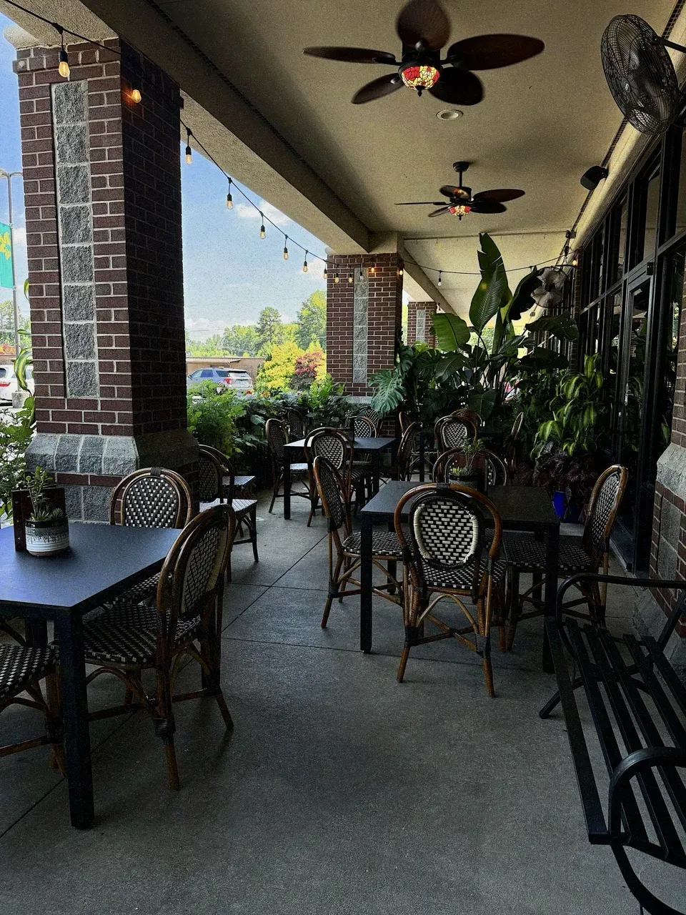 Covered patio with bistro chairs, lush plants, ceiling fans, and string lights